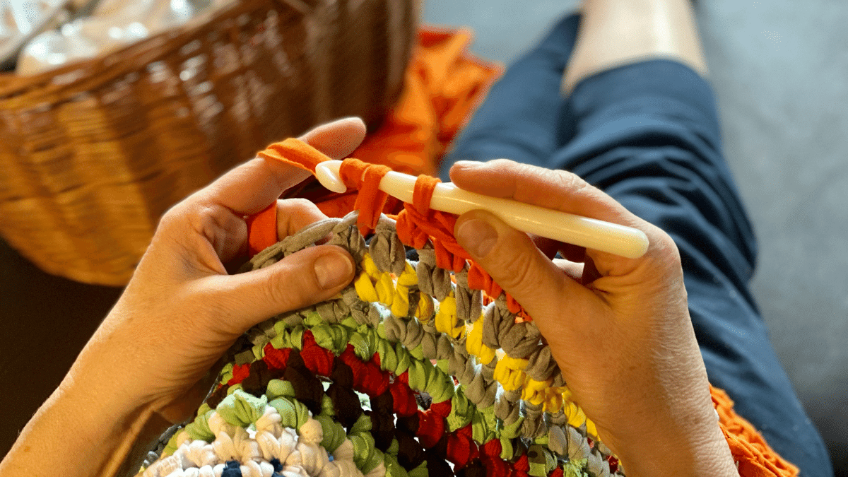close up of woman sitting down beside a basket crocheting a multi coloured pattern